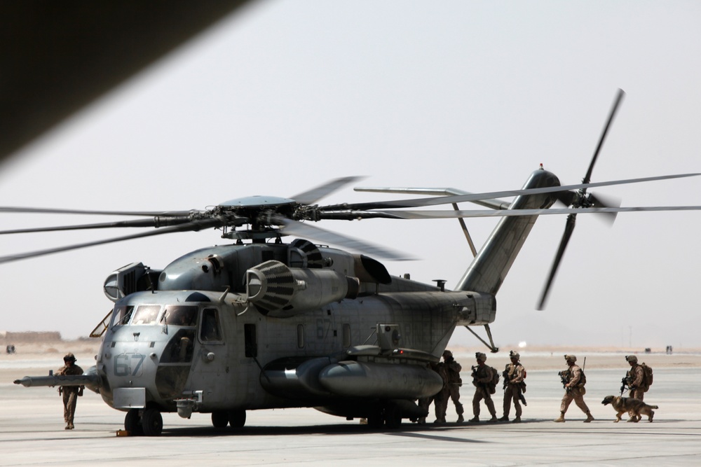 A large military heavy-lift helicopter, marked with the number '67', is on a tarmac in what appears to be an arid desert environment. Several military personnel in combat gear are gathered near the aircraft, and at least one military working dog is visible. The rotors appear to be spinning, suggesting the helicopter is either preparing for departure or has just landed.