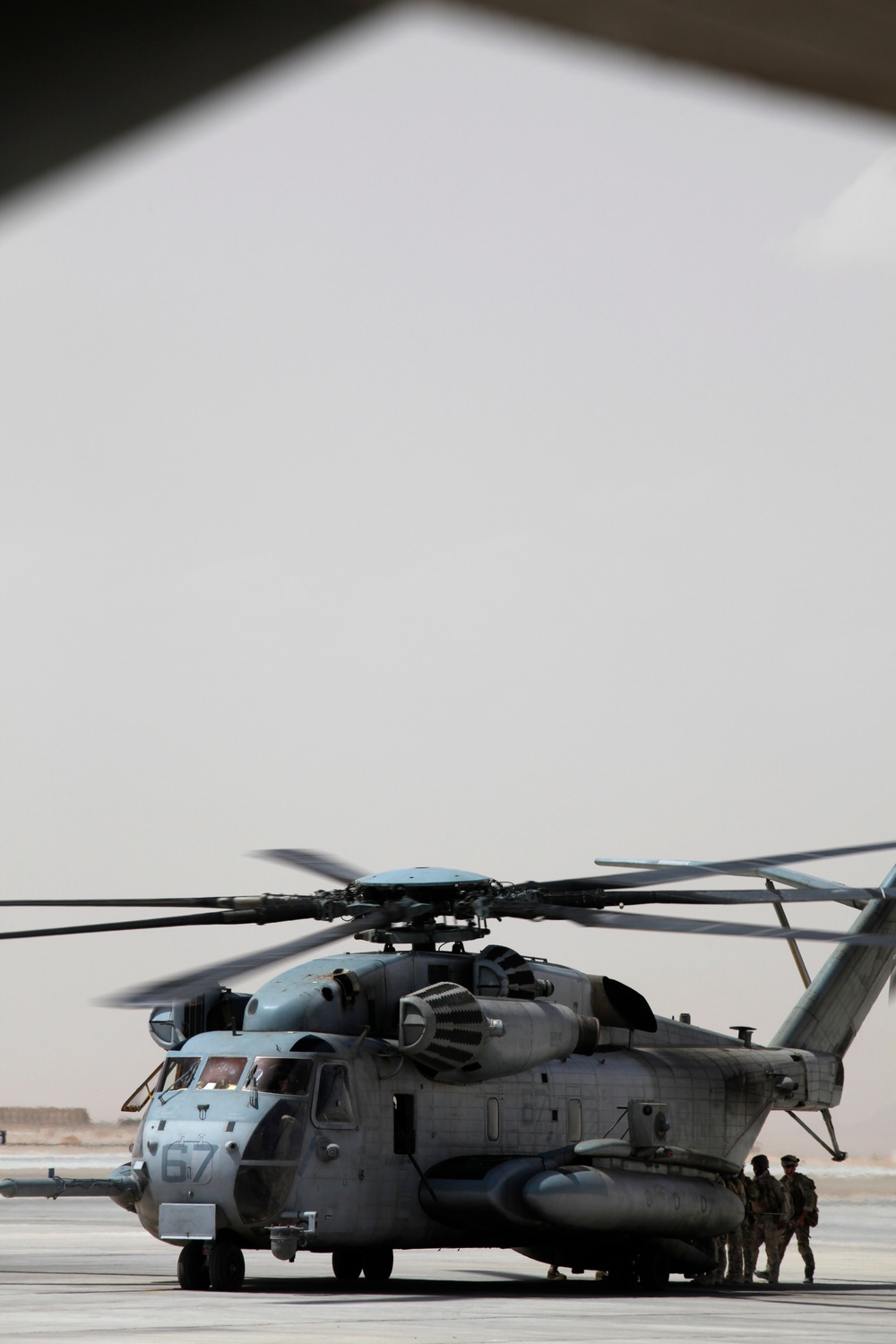 A large military heavy-lift helicopter, identifiable as a CH-53E Super Stallion bearing the number '67', is parked on a tarmac in what appears to be an arid desert environment. The rotors appear to be spinning or recently active, and several personnel in military gear are visible near the aircraft. The image is taken from beneath a structure, framing the scene through a triangular opening.