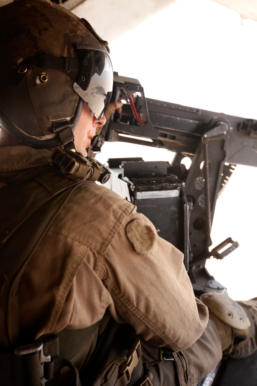 A military service member in full combat gear is positioned at an open door of a helicopter, looking outward. The individual is wearing a combat helmet with a mounted visor, tactical vest, and desert-tan uniform consistent with U.S. Marine Corps or Army attire. The setting appears to be an active aerial operation, likely in an arid or desert environment based on the dust-haze lighting.