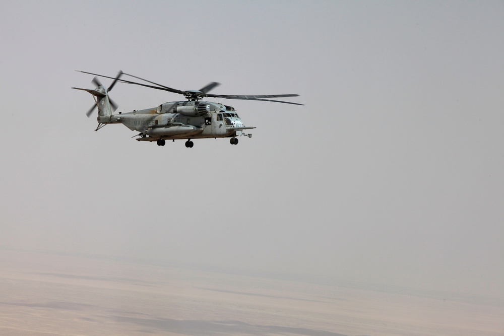 A large military helicopter, identifiable as a U.S. Marines CH-53E Super Stallion, is captured in flight over an arid desert landscape. The aircraft is flying at low-to-medium altitude with its rotor blades in motion, visible as a blur. The background shows a hazy, featureless desert terrain under an overcast sky.