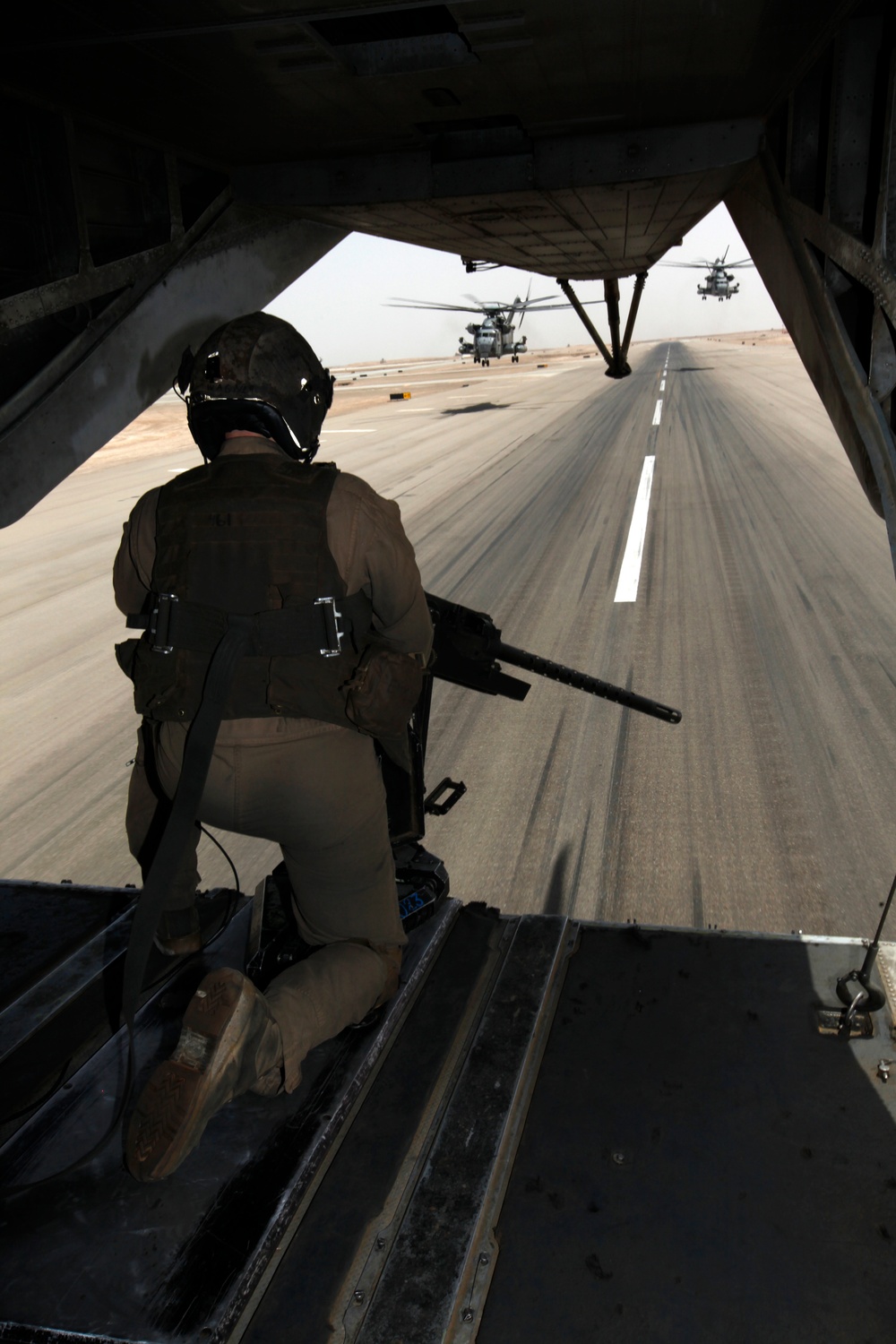 A military crew member is positioned at the open rear ramp of a helicopter, kneeling with a mounted weapon while the aircraft taxis or takes off along a runway. Two large military helicopters, identifiable as CH-53-type heavy-lift helicopters, are visible ahead on the tarmac in an arid environment. The motion blur on the runway surface indicates the aircraft is moving at speed.