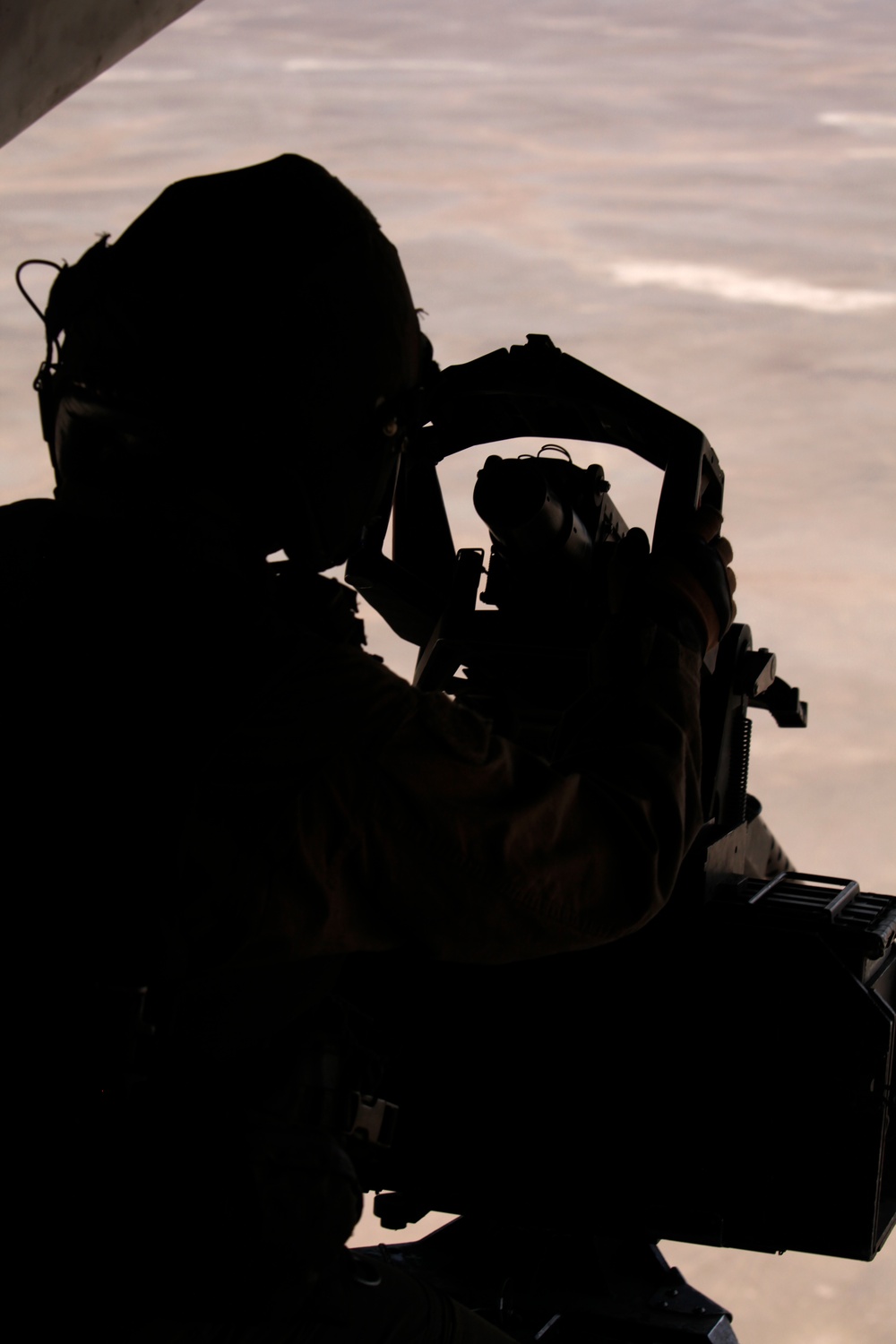 A silhouetted figure is positioned at what appears to be an open aircraft door or hatch, aiming a large crew-served or mounted weapon outward. The background reveals an arid, desert-like terrain viewed from altitude, suggesting this is an aerial platform. The strong backlight creates a complete silhouette, obscuring all identifying details of the person and equipment.