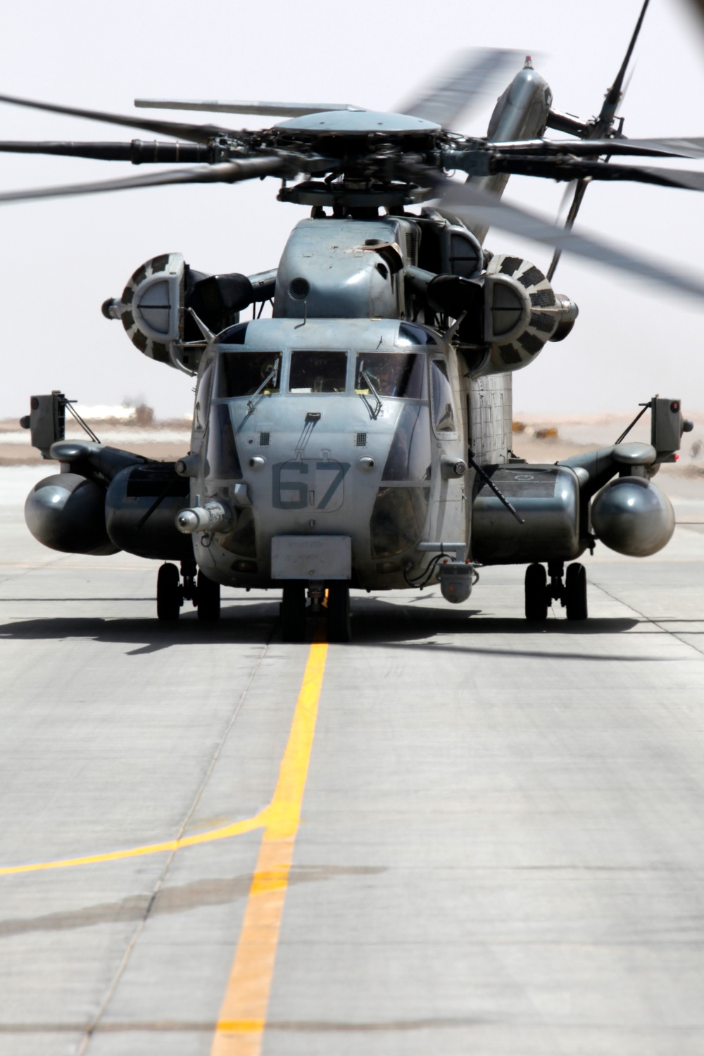 A large military helicopter, identifiable as a Sikorsky CH-53E Super Stallion, is photographed head-on while parked on a concrete flight line or tarmac. The aircraft bears the modex number '67' on its nose and is painted in standard military gray. The rotor blades are spinning, as evidenced by motion blur visible at the top of the frame.