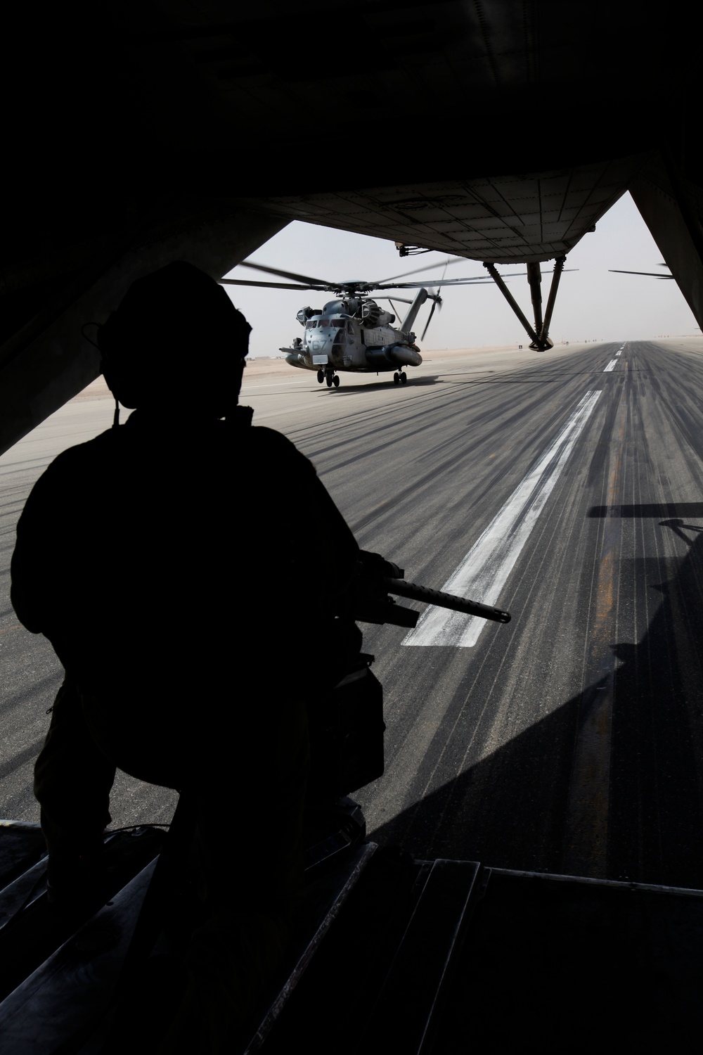 A silhouetted military personnel stands at the open rear ramp of a large military transport aircraft, looking out onto a tarmac where a heavy-lift helicopter is parked. The scene appears to be at a desert airfield, suggested by the arid, hazy horizon. The dramatic backlighting creates a strong silhouette contrast between the dark interior and the bright exterior.