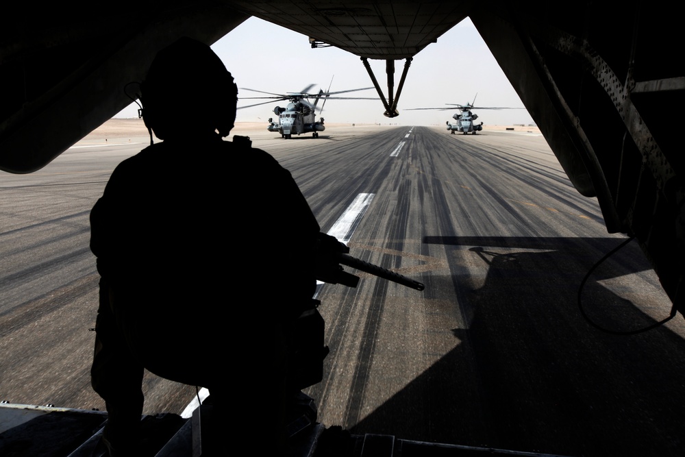 A silhouetted armed military personnel is observed from the open rear ramp of a large military transport aircraft, looking out onto a tarmac where two large military helicopters are positioned. The scene appears to take place at a desert airfield, suggested by the arid, hazy horizon. The composition creates a dramatic framing effect using the aircraft's ramp structure.