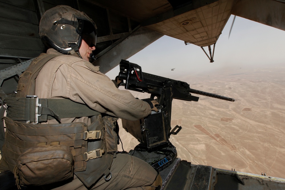 A military door gunner is positioned at the open door of a helicopter in flight over an arid desert landscape. The gunner is manning a crew-served weapon system mounted on a swivel mount, wearing full flight gear and tactical equipment. A second helicopter is visible in the background through the hazy desert atmosphere.