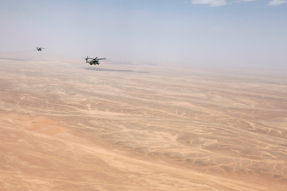 Two military helicopters are flying in formation over a vast, arid desert landscape. The terrain below is flat and sandy with minimal vegetation, consistent with a Middle Eastern or Central Asian desert environment. The image is taken from an aerial perspective, likely from a third aircraft flying alongside the formation.
