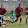 A rugby match in progress on a grass field, with players from two teams actively engaged in play. The red-and-navy team appears to have possession, with one player carrying the ball while teammates provide support. Several opposing players in teal/green uniforms are on the ground following a tackle.