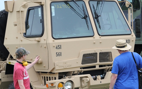 Two civilians are examining a tan/desert-camouflage military truck, likely at a public military vehicle display or air show. The large tactical truck bears identification numbers 451, 561, and 732905, and appears to be an LMTV or FMTV-type Army vehicle. A second camouflage-painted military truck is partially visible on the right side of the frame.