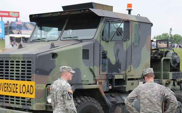 A military equipment display at what appears to be a public event or air show, featuring a large camouflage-painted military heavy transport truck with an 'OVERSIZE LOAD' sign on its front bumper. Two U.S. Army soldiers in ACU (Army Combat Uniform) are speaking with civilian visitors on a grass field. A sign in the background reads 'Blaine Johnson Grandstand' and 'Stormont-Vail HealthCare', suggesting a fairground or event venue.