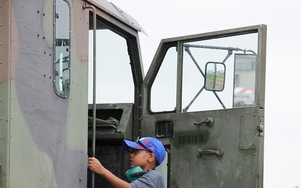 A young boy wearing a blue cap and casual clothing is climbing up the side of a large military tactical truck, gripping a handle for support. A U.S. Army soldier in ACU (Army Combat Uniform) pattern camouflage stands nearby, watching the boy and shielding his eyes with one hand. The setting appears to be an outdoor military display or public event on a grassy area.