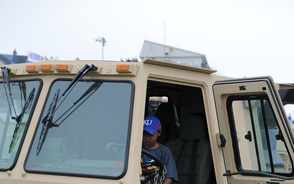 A U.S. Army soldier in ACU (Army Combat Uniform) assists a young boy wearing a KU (Kansas University) baseball cap who is sitting in the cab of a military tactical truck. The vehicle is a tan/desert-colored heavy military truck marked with the identifiers '821', 'HQ60', and 'C 05'. The scene appears to be a public military display or community outreach event held on a grass field.