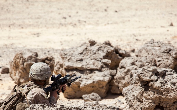A soldier in desert camouflage uniform is positioned behind rocky cover in an arid, desert environment, aiming a rifle equipped with an optical scope. The soldier appears to be in a prone or crouched firing position using the natural rock formation as concealment. The setting is consistent with a desert training exercise or operational deployment.