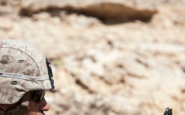 A U.S. Marine in desert MARPAT camouflage is positioned behind rocky terrain in an arid environment, holding an M4 carbine rifle equipped with an optic scope and underbarrel attachment. The soldier appears to be in a prone or low-profile defensive/observation position. A name tape reading 'TORRES' is visible on the helmet band.