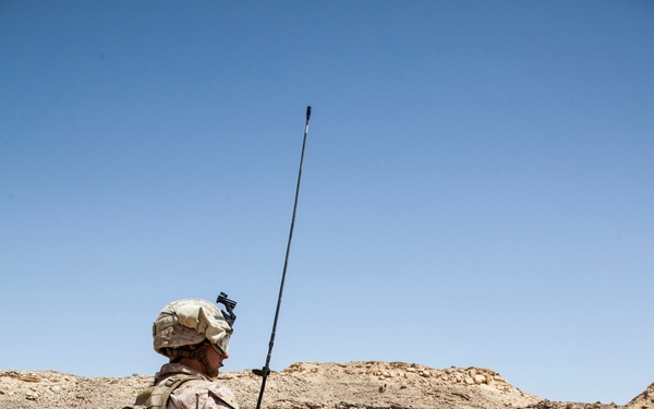 Three military personnel in desert camouflage uniforms are positioned in a rocky, arid desert terrain. The foreground soldier is standing with one foot elevated on a rock and carries a radio with a tall whip antenna extending above. The other two soldiers appear to be in kneeling or crouching positions, suggesting a tactical observation or patrol halt.