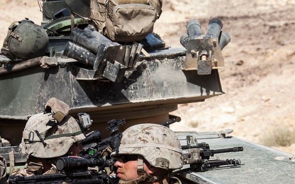Two military personnel in desert camouflage uniforms are positioned on an armored vehicle, aiming rifles in a desert environment. The soldiers are equipped with full combat gear including helmets, body armor, and optical sights on their weapons. The scene appears to be a combat operation or training exercise in an arid, rocky terrain.