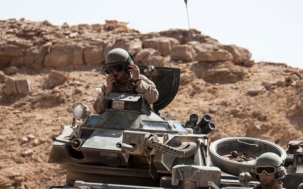 The image shows two military personnel operating armored vehicles in an arid, rocky desert environment. One soldier is visible in an elevated hatch position using communication equipment, while a second soldier is partially visible in a separate hatch wearing protective goggles. The vehicles appear to be amphibious assault vehicles or armored personnel carriers moving through rugged terrain.