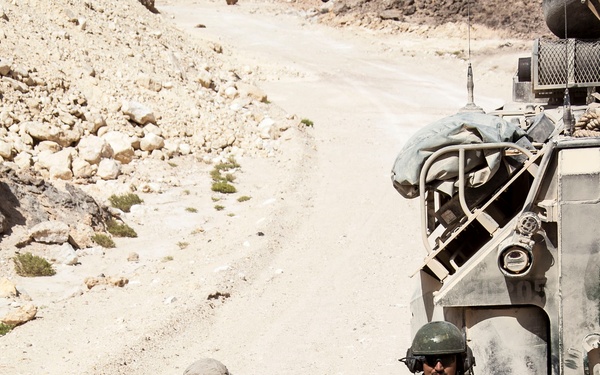 Four military personnel in desert camouflage uniforms are gathered near a military armored vehicle on a dirt road in an arid, rocky terrain. The soldiers appear to be coordinating or assisting one another, possibly during a patrol or operational movement. The setting suggests a desert environment consistent with Middle Eastern or Central Asian terrain.