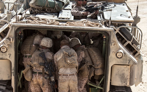 The image shows military personnel in desert camouflage uniforms loading or unloading from the rear ramp of a tracked armored personnel carrier (APC). The vehicle appears to be a LAV or similar wheeled/tracked military transport, painted in desert tan, operating in an arid, rocky terrain. Additional personnel are visible on top of the vehicle, one appearing to man a weapon system.