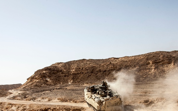 A tracked armored personnel carrier (APC) is moving at speed across an arid desert terrain, kicking up a large dust cloud behind it. The vehicle appears to be desert-camouflaged and has personnel visible in the open hatch on top. The landscape consists of rocky, barren hills with a dirt track winding through the terrain.