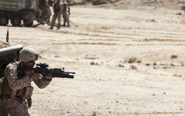 A soldier in desert camouflage uniform is exiting or taking position from the rear ramp of an armored personnel carrier (APC) while aiming a rifle with an underbarrel grenade launcher attachment. The scene takes place in an arid, desert environment with sparse vegetation. Additional military personnel and a large wheeled military vehicle are visible in the background.
