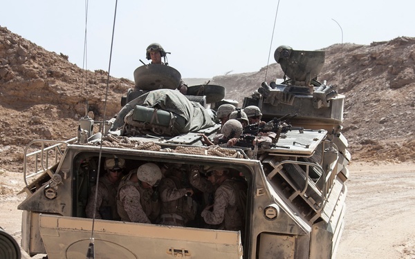 The image shows military personnel riding in what appears to be an AAV-7 (Amphibious Assault Vehicle) moving along a dirt road in an arid, rocky desert environment. Multiple soldiers are visible both inside the open rear troop compartment and on top of the vehicle, with gear and equipment strapped to the exterior. The photo is taken from the perspective of a following vehicle in the same convoy.