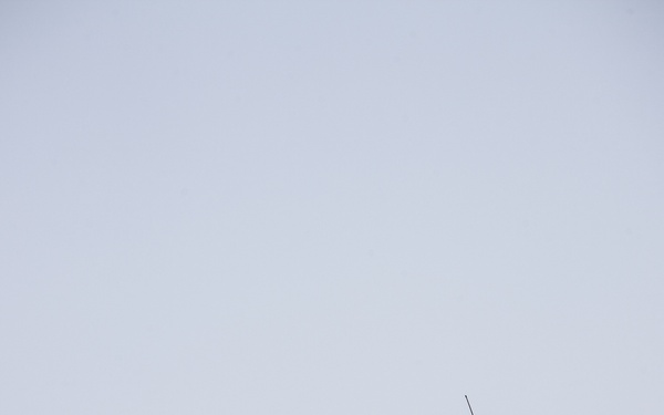 A single soldier in desert camouflage stands atop a rocky ridge or hilltop against a clear, pale blue sky. The individual appears to be holding a radio or communication device with an antenna raised above their head, likely signaling or communicating. The arid, rocky terrain and bright lighting suggest a desert environment.