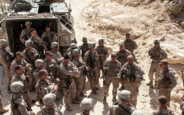 A large group of military personnel in desert camouflage uniforms are gathered in a briefing or debriefing formation in an arid, rocky terrain. The soldiers are assembled around what appears to be a central figure who may be addressing the group, with a military tactical vehicle visible in the background. The scene suggests a pre- or post-mission briefing in a desert environment.