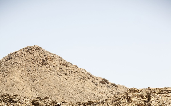 The image shows a small group of military personnel in desert camouflage uniforms ascending a rocky, arid hillside. The terrain is characteristic of a desert environment with layered sedimentary rock formations and sparse vegetation. The soldiers appear to be conducting a patrol or reconnaissance operation in a harsh, sun-baked landscape.
