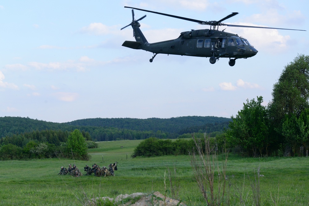 A military helicopter, identifiable as a UH-60 Black Hawk, hovers low over an open green field while a group of soldiers in combat gear assembles or takes cover on the ground below. The scene appears to depict a tactical insertion or extraction exercise in a rural, forested landscape. The setting suggests a military training operation conducted in a temperate European-style environment.