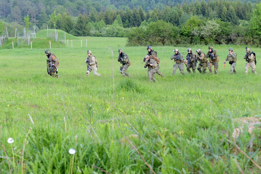 A group of approximately 9-10 armed military personnel in full combat gear are moving tactically across an open green field. The soldiers appear to be conducting a training exercise, advancing in a loose formation from right to left across the grassy terrain. The setting includes a treeline in the background and what appears to be a military training range with marker poles visible.