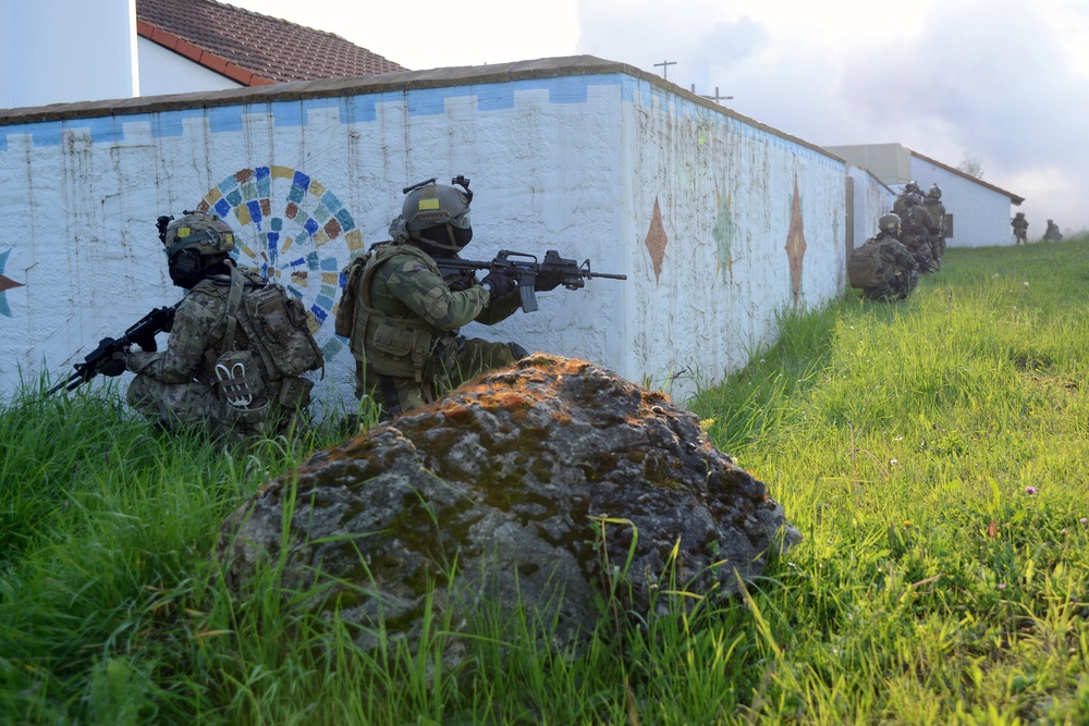 The image shows a group of heavily equipped military or special operations personnel conducting what appears to be a tactical training exercise along a decorated perimeter wall. The soldiers are in full combat gear including helmets, body armor, and carrying assault rifles, moving in a tactical formation along a grassy slope. The setting appears to be a training facility or urban environment with a colorful mural-decorated white wall.