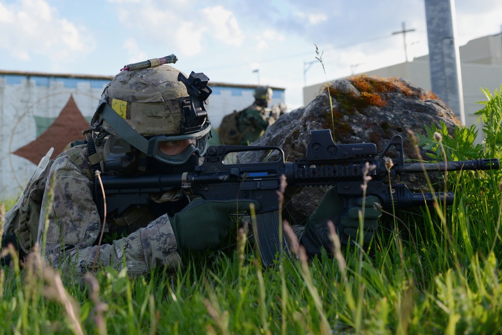 A soldier in full combat gear is prone in tall grass, aiming an assault rifle equipped with a red dot sight. A second soldier is visible in the background near a large rock. The setting appears to be a military training exercise in an urban-adjacent environment.