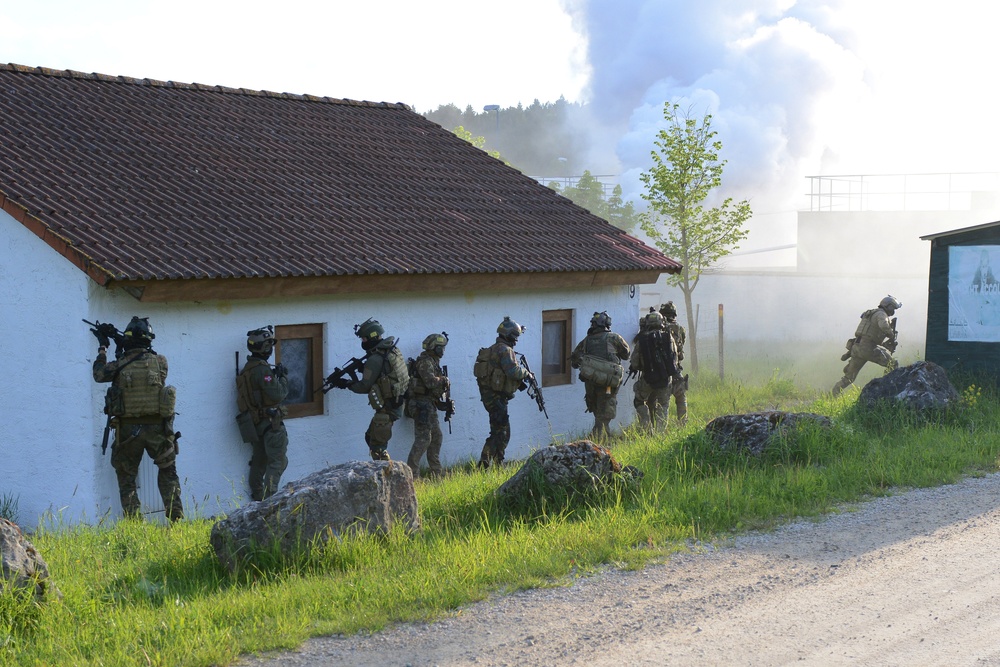A group of heavily armed and equipped military or special operations personnel are conducting a tactical building clearance exercise along the exterior wall of a white stucco building with a red tile roof. The personnel are moving in a stacked formation, suggesting a coordinated entry or assault drill. Smoke is visible in the background, consistent with a training simulation environment.