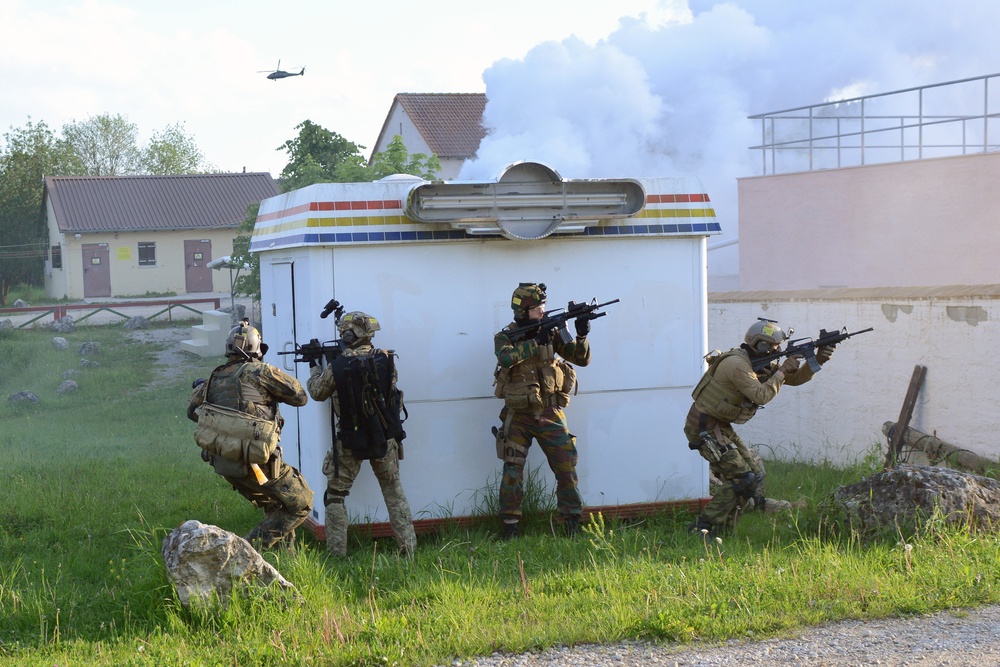 The image depicts a military tactical training exercise with four armed soldiers in combat gear taking defensive positions around a white trailer or kiosk structure. White smoke is visible in the background, and a helicopter can be seen in the sky, suggesting a coordinated multi-element drill. The setting appears to be a mock village or urban training environment.
