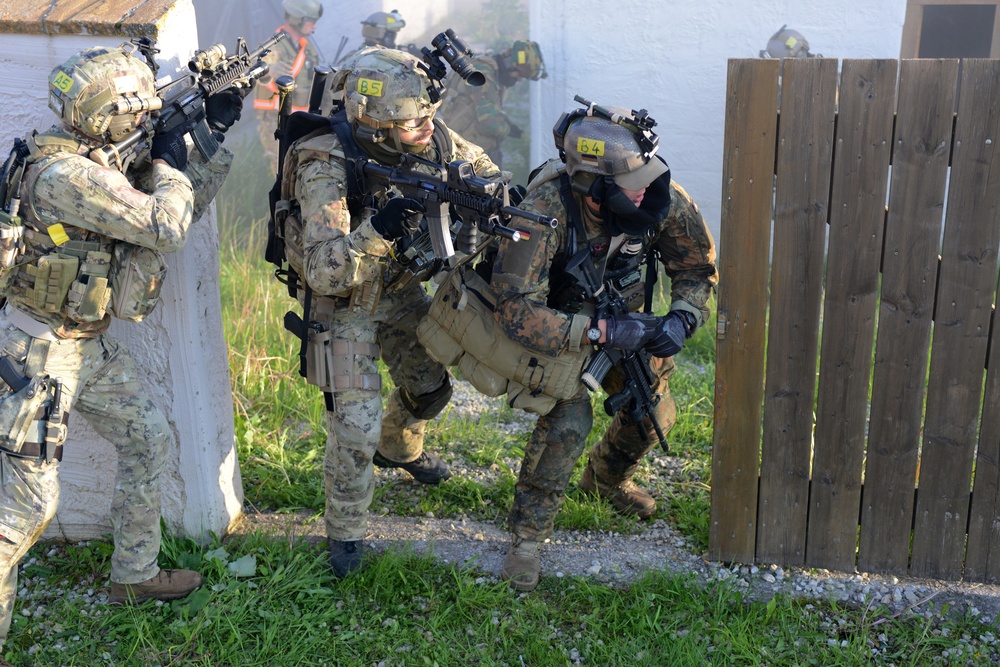 The image shows a group of heavily armed military or special operations personnel conducting what appears to be a tactical training exercise in an urban environment. Multiple soldiers in mixed camouflage patterns are moving in formation near a building and wooden fence structure. Yellow numbered identification tags on their helmets suggest this is a coordinated exercise or drill rather than a combat operation.