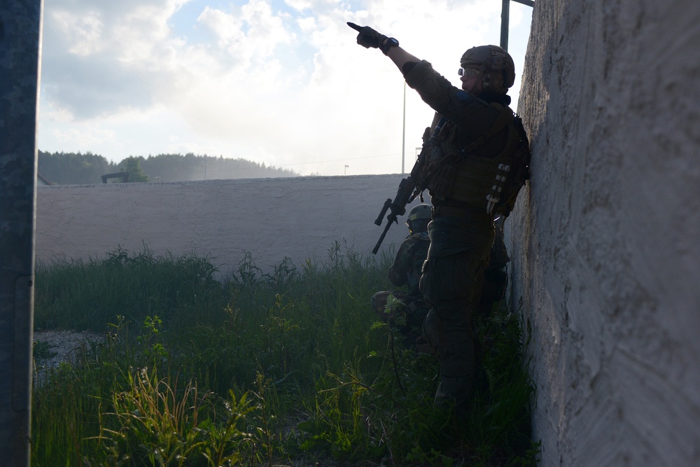 Two armed military or tactical personnel are positioned against a concrete wall in what appears to be a training exercise or tactical operation. The lead operator is pointing upward/forward while holding a rifle, with a second operator crouched behind. The setting includes overgrown vegetation, a concrete barrier wall, and a hazy background suggesting possible smoke or dust.