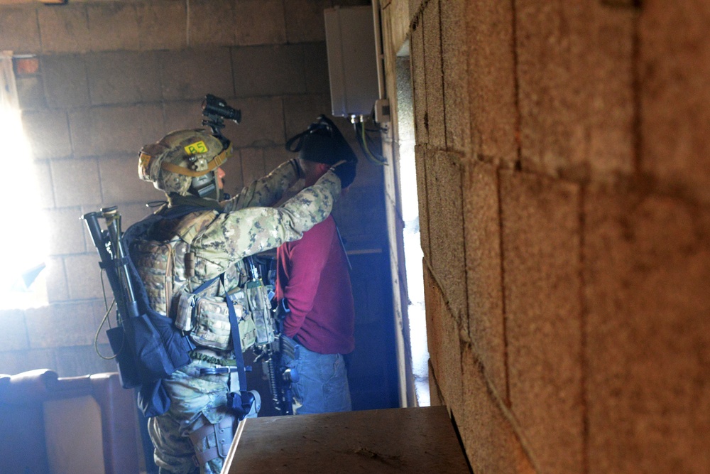 A fully equipped military or law enforcement operator in multicam camouflage and tactical gear is conducting what appears to be a room-clearing or detainee-control exercise inside a concrete block structure. The operator is physically engaging with a second individual wearing civilian clothing (red shirt, jeans). The setting and yellow marker on the helmet suggest this may be a training exercise.
