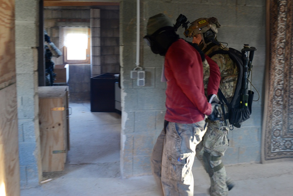 The image shows a military or law enforcement training exercise taking place inside a concrete block structure. A soldier in full tactical gear and camouflage is escorting or detaining a role-player wearing a red sweatshirt with a dark hood/balaclava covering their face. A second armed figure is visible in the background room, suggesting a coordinated room-clearing or detainee-handling drill.