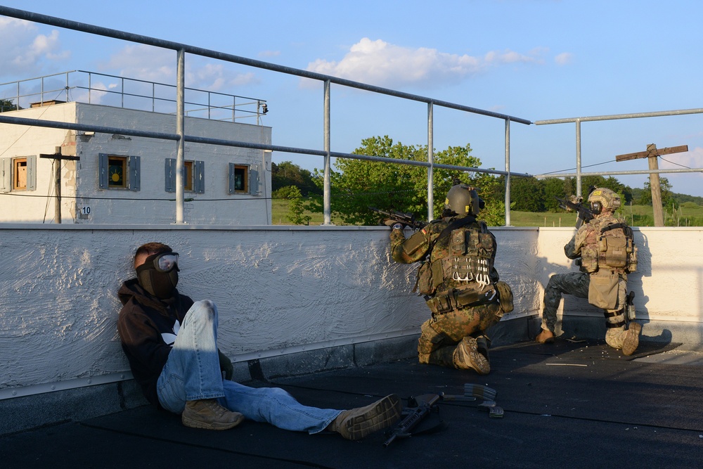 The image depicts what appears to be a military or law enforcement training exercise on a rooftop. Two armed personnel in tactical gear and helmets are positioned near a low wall, with one individual seated against the wall wearing civilian clothing and protective goggles, likely playing the role of a detainee or hostage. The setting includes a training facility building visible in the background with target markers on its windows.