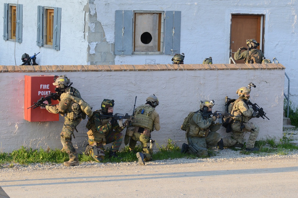 The image depicts a military tactical training exercise taking place at what appears to be a mock urban environment or training facility. Multiple armed soldiers in full combat gear are positioned along a white perimeter wall, taking cover and aiming weapons in a coordinated assault formation. Additional personnel are visible on the upper level of the building, providing overwatch positions.