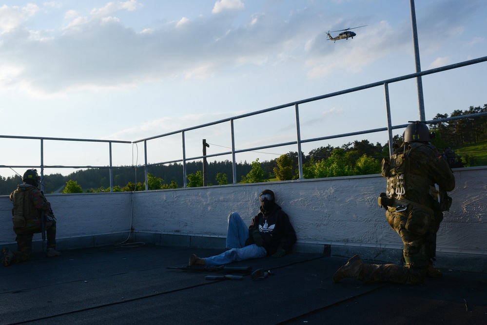 The image depicts what appears to be a military or law enforcement training exercise on a rooftop. Two armed personnel in tactical gear and helmets are positioned around a seated civilian role-player wearing goggles, a black Adidas hoodie, and jeans. A military helicopter, consistent with a UH-60 Black Hawk, is visible in the background sky.