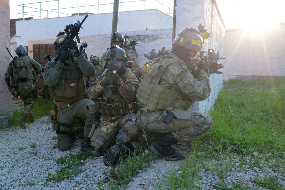 A group of heavily armed and equipped military or special operations personnel are conducting what appears to be a tactical training exercise in an urban environment. The operators are positioned along a building wall in various tactical stances, with weapons raised and aimed. The scene is illuminated by a bright sun flare from the right side, suggesting early morning or late afternoon timing.