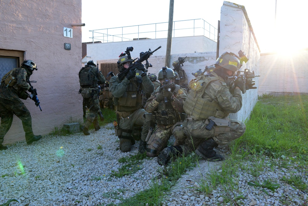 A group of heavily armed and equipped tactical/military personnel are conducting what appears to be a training exercise in an urban environment. The personnel are wearing mixed camouflage patterns and full tactical gear, taking up positions along a wall near a building numbered '11'. The scene is backlit by a low sun, suggesting early morning or late afternoon conditions.