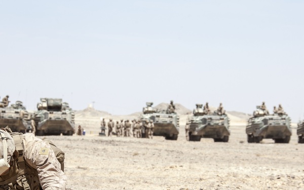 A military exercise or operation in a desert environment showing soldiers in desert camouflage uniforms in the foreground, with a large formation of armored vehicles lined up in the background. A soldier is crouching down handling what appears to be an ammunition can or equipment case. The scene suggests a large-scale military exercise or staging operation.