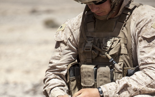 A U.S. Marine in desert MARPAT camouflage and tactical vest is kneeling on arid, rocky terrain while handling a wire or cord connected to a green military equipment box on the ground. The soldier appears to be conducting a training or operational exercise involving what appears to be a demolitions or communications device. The setting is a dry, desert environment consistent with a military training range.