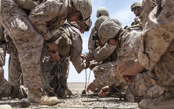 A group of military personnel in desert camouflage uniforms are gathered in an arid, rocky environment working with cables or wiring on the ground. The soldiers appear to be conducting a training exercise or field operation involving what appears to be detonation cord or communication cable assembly. The setting is a barren desert landscape under clear skies.