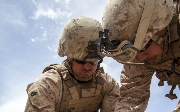 Two military personnel in desert camouflage uniforms are kneeling on arid, rocky ground while handling what appears to be detonation cord or wire. Both wear MARPAT desert pattern uniforms, tactical vests, and helmets with night vision goggle mounts, consistent with U.S. Marine Corps equipment. The activity appears to be related to explosive ordnance or demolition training/operations.