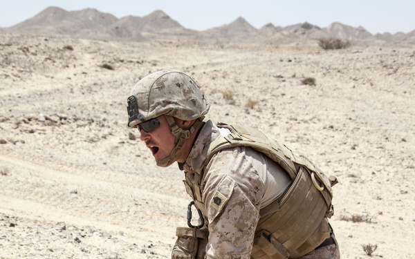 A soldier in desert camouflage uniform is crouched forward in an arid desert environment, pulling a cord or wire while shouting. The setting appears to be a training exercise or operational activity in a rocky desert landscape with mountains visible in the background. The soldier is fully equipped with body armor, helmet, tactical gloves, and a backpack.
