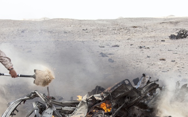 A military service member in desert camouflage uniform is attempting to extinguish or manage a burning, heavily destroyed vehicle in an arid desert environment. The vehicle appears to have been destroyed by an explosion, with wreckage scattered across the sandy terrain. Smoke and active flames are visible amid the twisted metal debris.